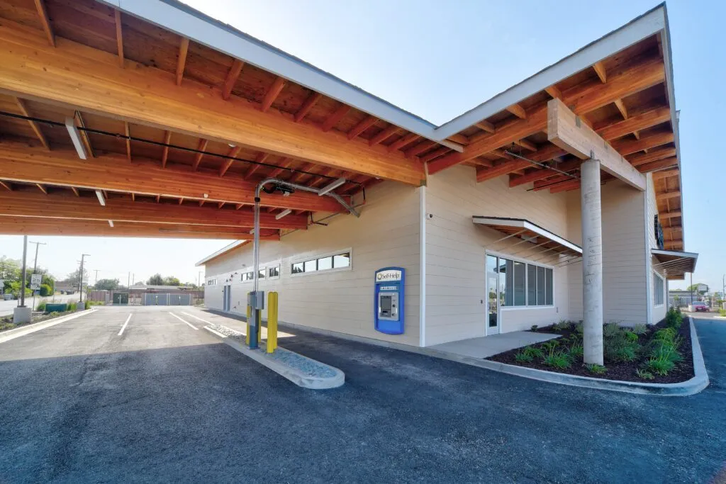 An exterior view of the Self-Help Federal Credit Union building's drive-thru, featuring a large wooden-paneled canopy supported by concrete columns, and an ATM.