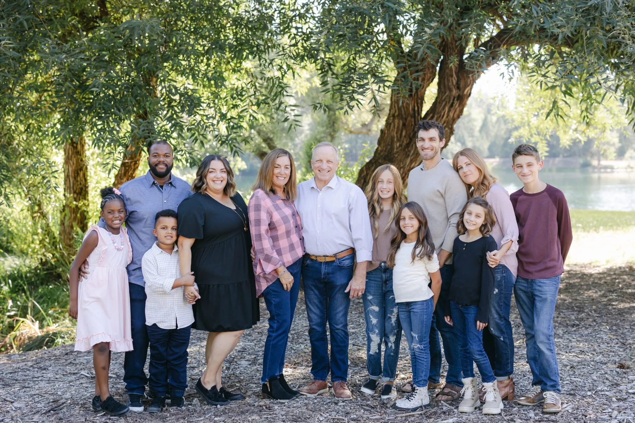 Guy and Gina Simile with their family, standing together beneath a tree beside a lake.